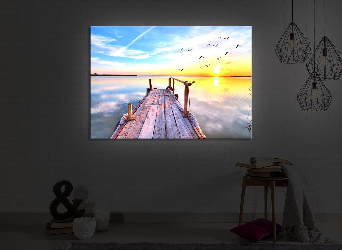 led bild mit dem Motiv Steg mit Möwen bei Sonnenuntergang in einem dunklen Raum. Das Bild leuchtet weiß im abgedunkelten Zimmer